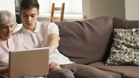 elderly woman and young man sitting on sofa in living room and talking while using laptop computer