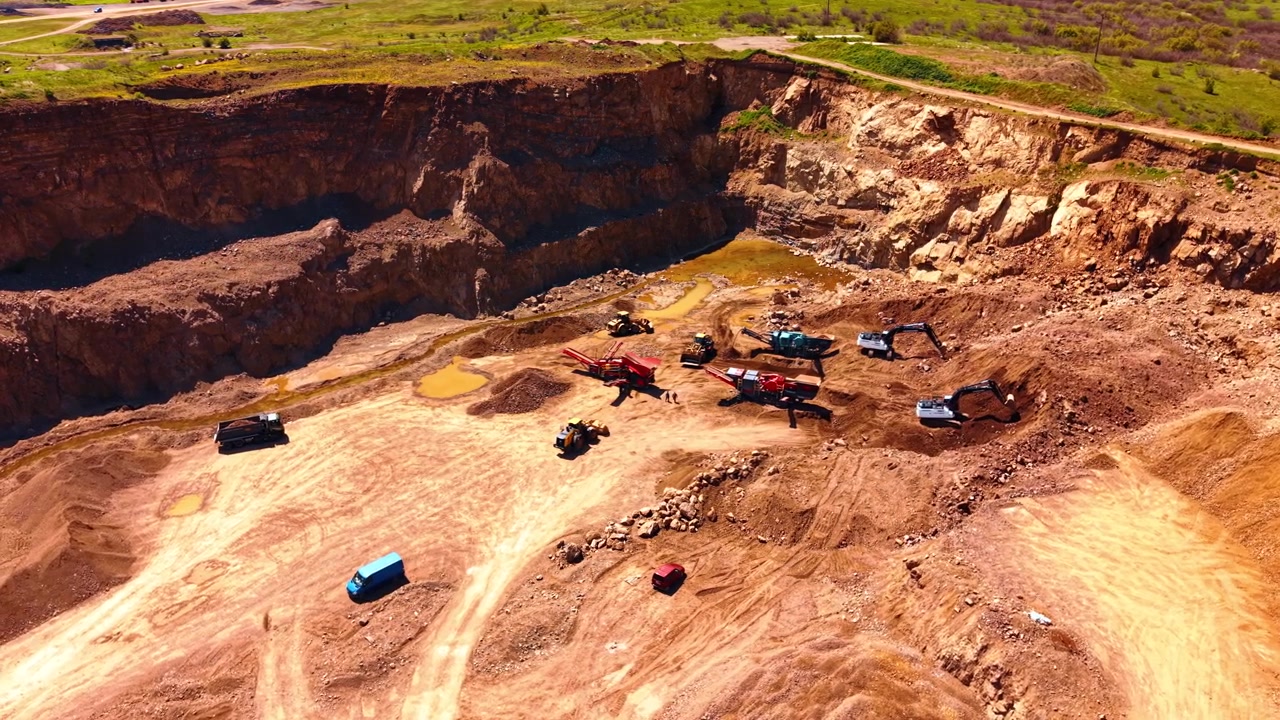 Open pit stone quarry with heavy machinery operating. Wide aerial view of excavators crushing plant and trucks operating inside active rock quarry pit.