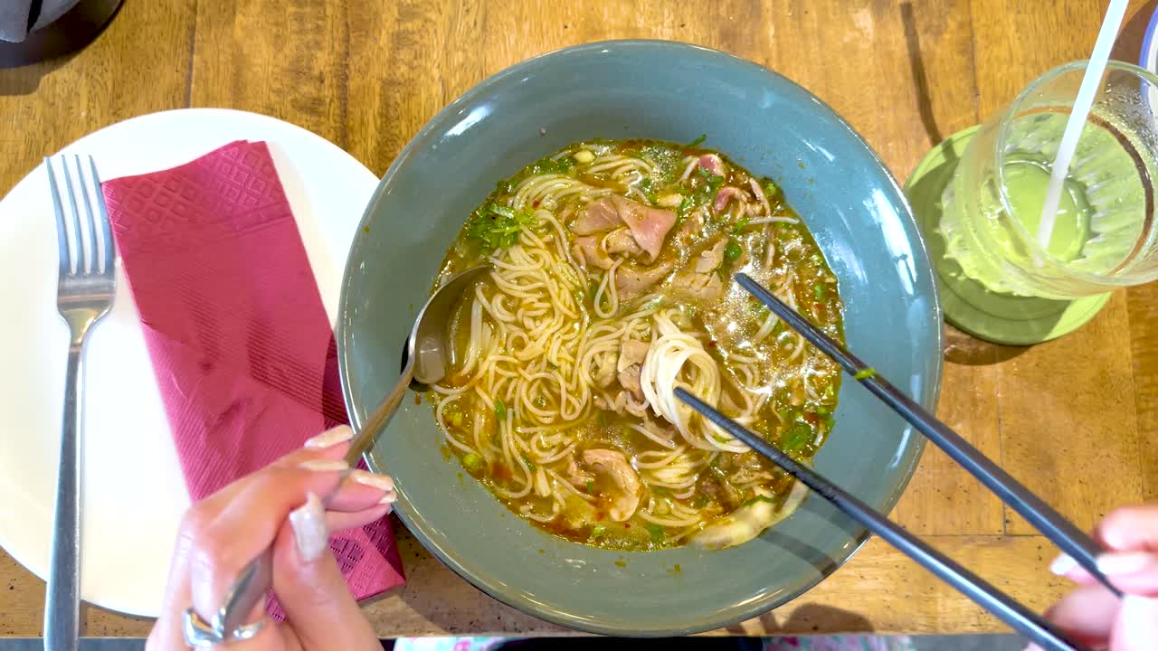 A person savors Vietnamese pho with chopsticks in a Bangkok eatery. Bright lighting highlights the dish's vibrant colors