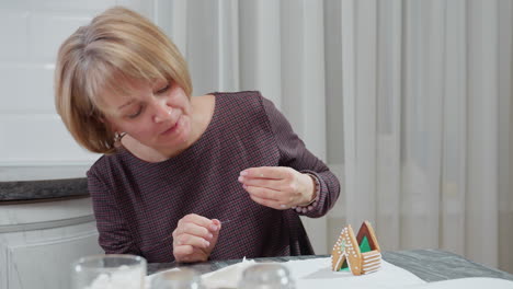 Young woman carefully decorating house-shaped cookie with icing while adjusting it, she licks icing and places it while other baking equipment and cookies are arranged on table