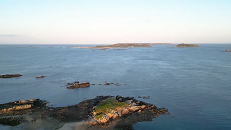 Dropping-aerial-view-from-Bordeaux-harbour-Guernsey-over-calm-waters-of-the-Little-Russell-Channel-to-Herm-and-Jethou-falling-to-reveal-dried-out-boats-in-late-afternoon-sun