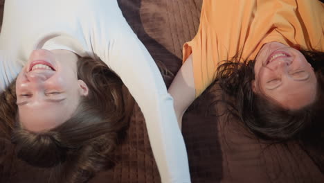 two cheerful ladies fall backward onto bed with joy and laughter in cozy wooden room, captured from behind showing spontaneous bonding moment and carefree energy