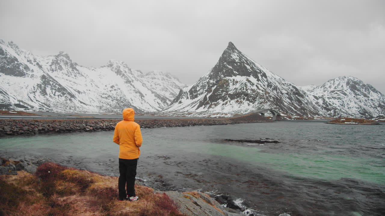 hombre con chaqueta amarilla caminando hacia el borde de la costa para admirar el hermoso paisaje invernal en noruega - plano amplio