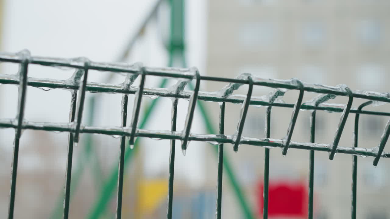 Close-up of a green metal fence coated in ice with icicles clinging to the wires, set against a blurred snowy playground background, capturing a serene yet chilly winter scene