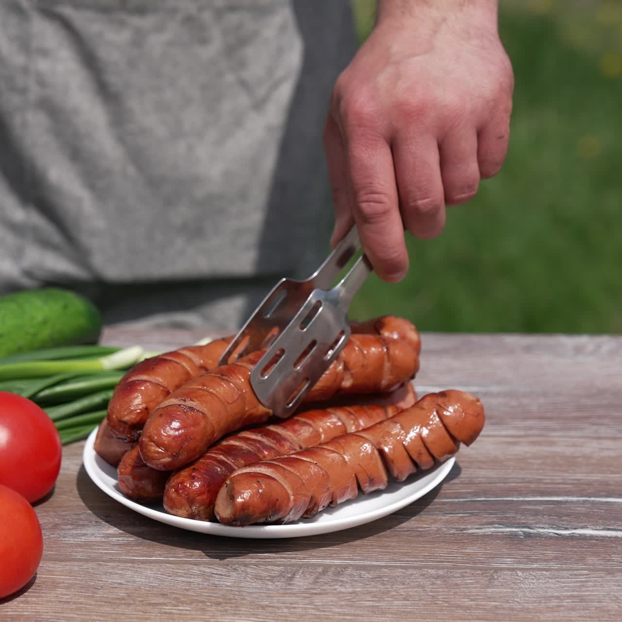 Ready grilling sausages with vegetables on the table outdoors. Chef puts roast sausage into a plate. Tasty meal for a picnic