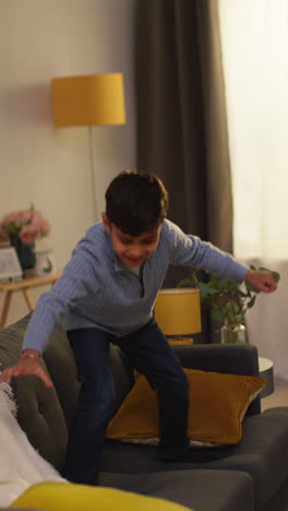 vertical video of disruptive young boy behaving badly at home jumping on sofa and throwing cushions around lounge 1