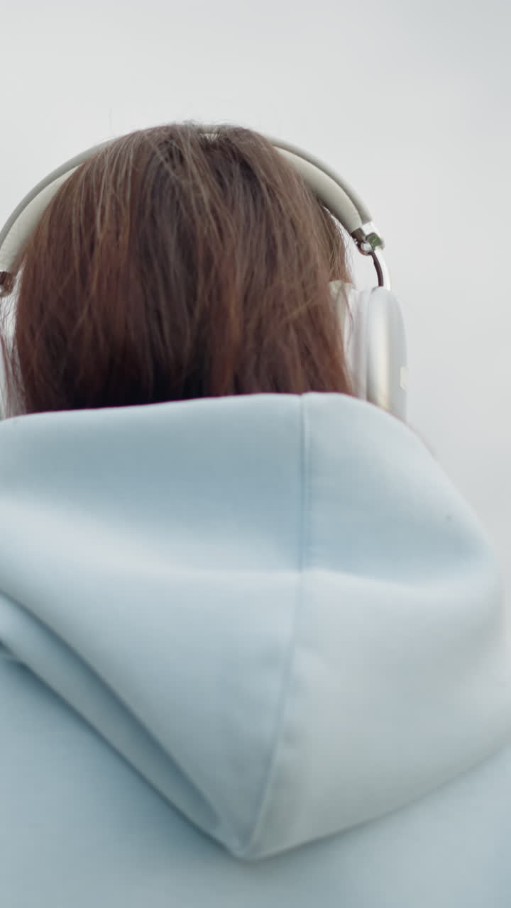 Back view of young lady in blue sweater and headphones walking in cool environment, blurred withered tree in background, adding a calm and serene vibe to the outdoor scene