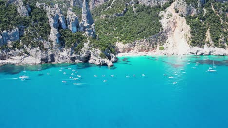tourist boats enjoy summer vacation at cala goloritze beach rock formations in sardinia, italy - aerial 4k tilting reveal