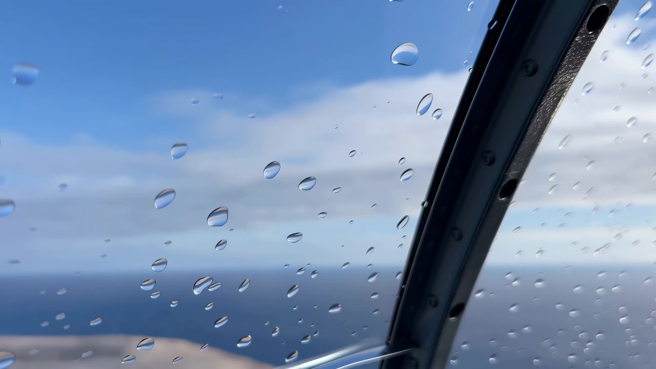 Clear droplets remain scattered across glass while pale clouds shift subtly beyond, creating layered focus between wet window surface, blue horizon, and dark structural frame.