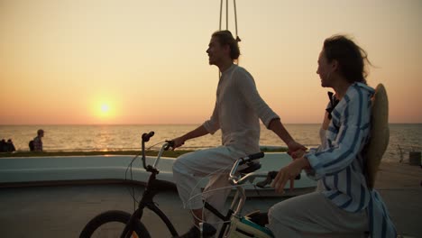 a guy in light summer clothes and a girl in a striped shirt ride bicycles along the sea at dawn. a guy and a girl are driving along the beach at dawn