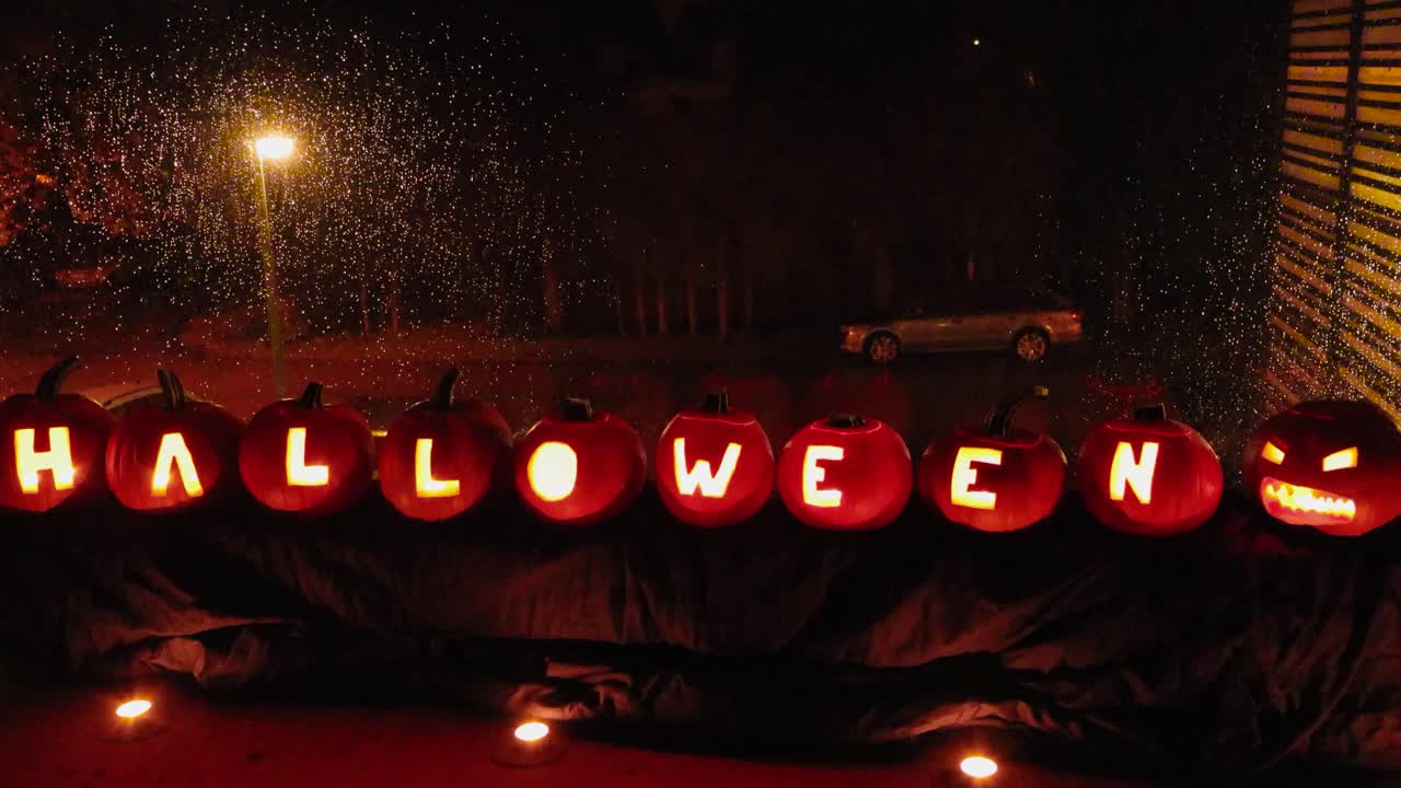 Carved out Halloween pumpkins that have glowing candle light in them in front of a window during autumn evening or night time while it's raining and raindrops are on window. Candle light reflecting.
