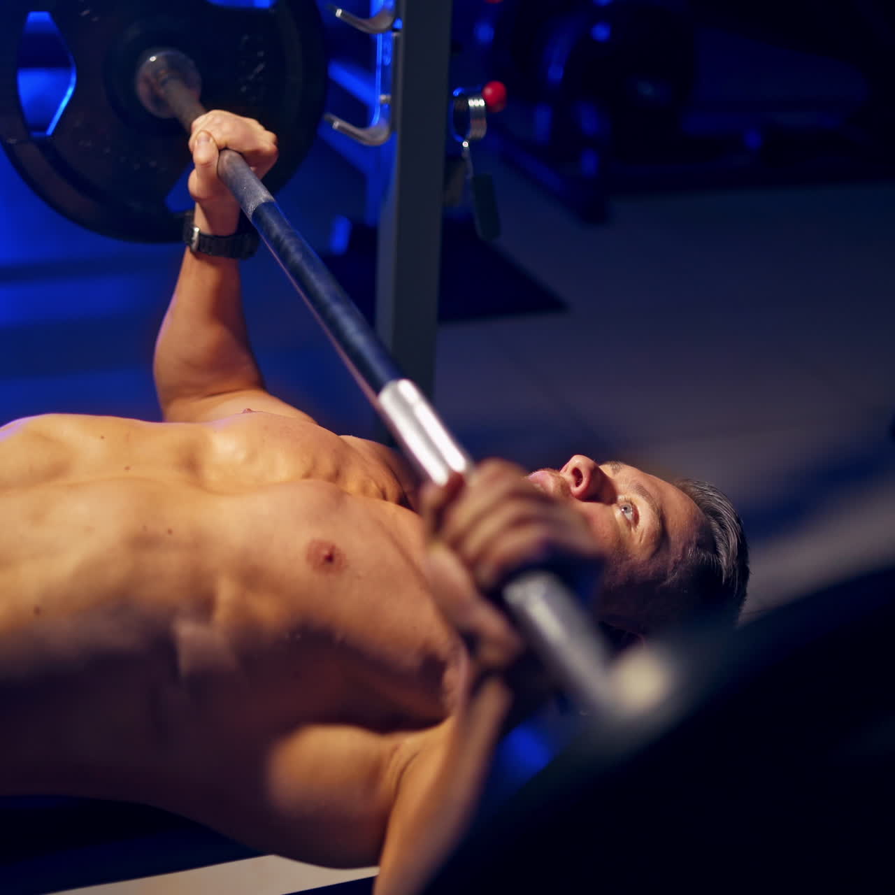Shirtless young bodybuilder doing bench press for chest. Strong abs. Blue light filter. Video from the side.