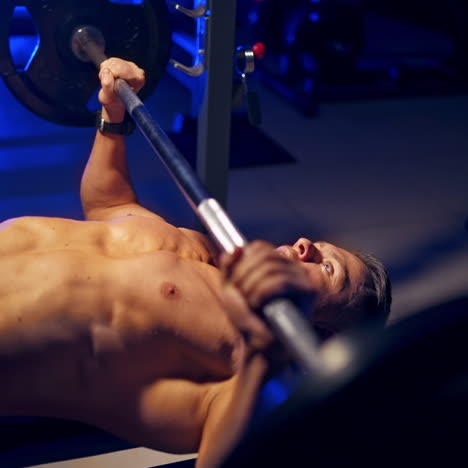 shirtless young bodybuilder doing bench press for chest. strong abs. blue light filter. video from the side.