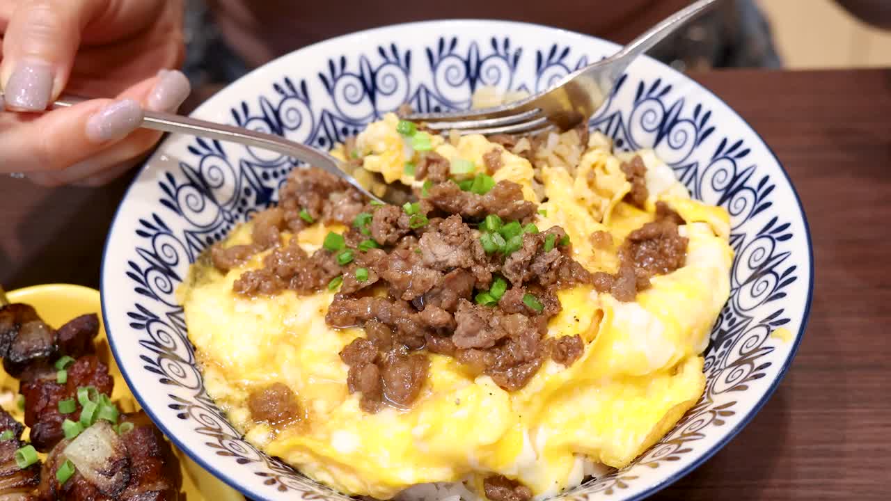 A person uses a spoon and fork to eat a bowl of Thai-style beef and egg rice in a brightly lit indoor setting, captured in close-up