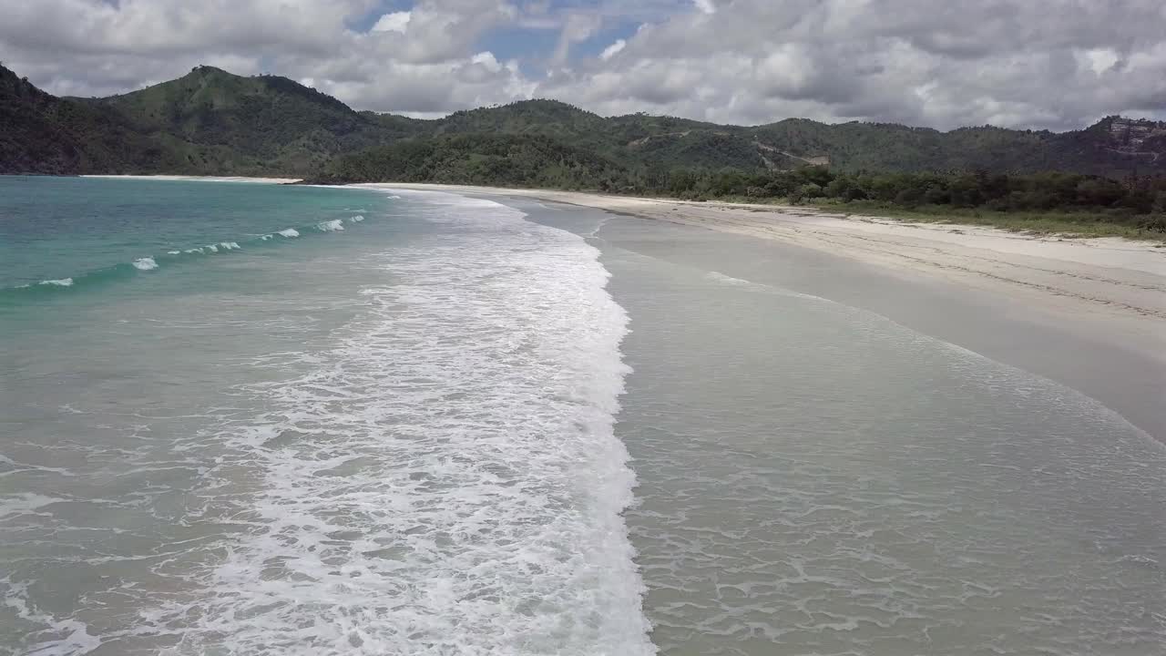 Aerial view of Selong beach at Lombok Island,white waves washing the white sand beach,surrounded by green mountains, Indonesia.Pan left towards the sea.