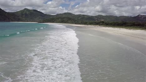 aerial view of selong beach at lombok island,white waves washing the white sand beach,surrounded by green mountains, indonesia.pan left towards the sea.