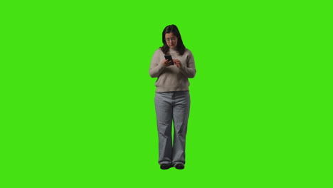 young woman sending or reading text message or scrolling online on mobile phone standing against green screen