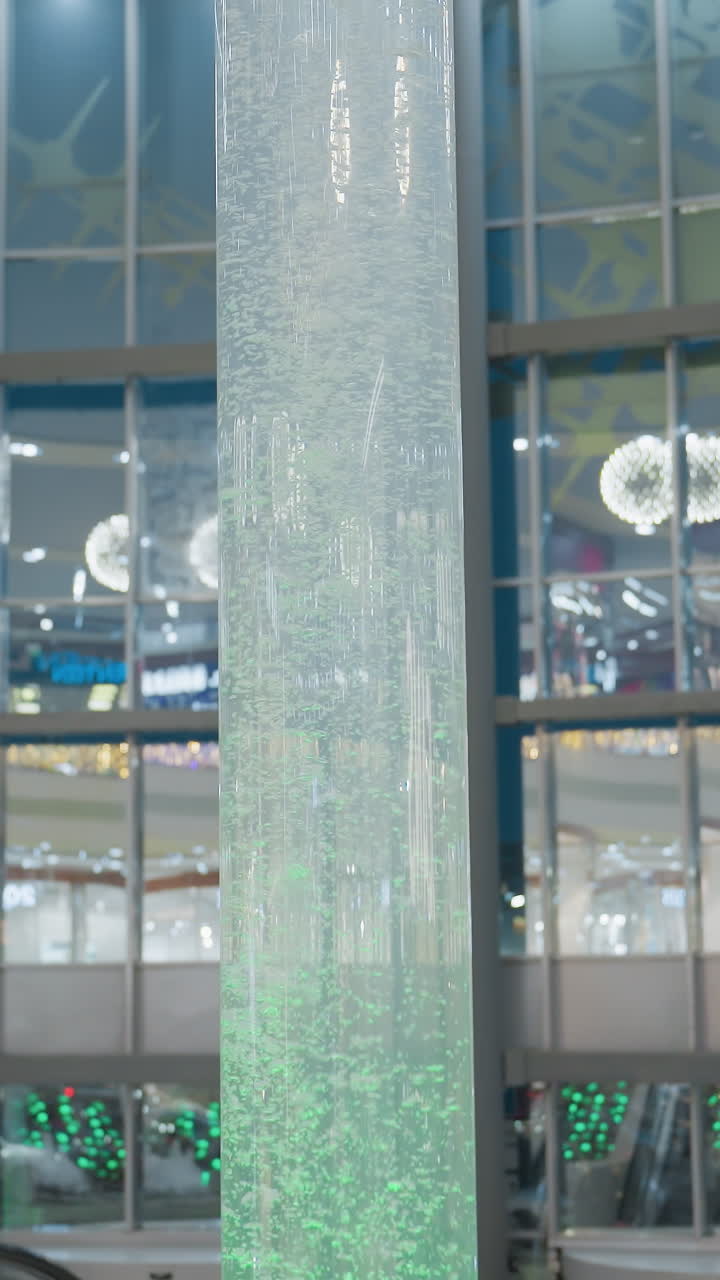 Beautiful Christmas decorations inside mall with glowing lights, festive ornaments hanging on trees, and large cylindrical water feature in center, creating warm and cozy holiday atmosphere
