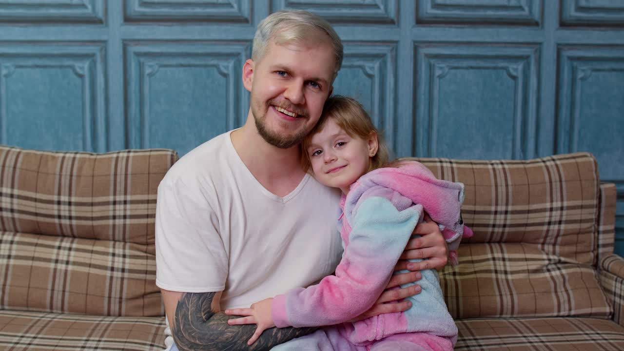 Father and little child daughter kid in pajamas sit on couch in room smiling, looking at camera