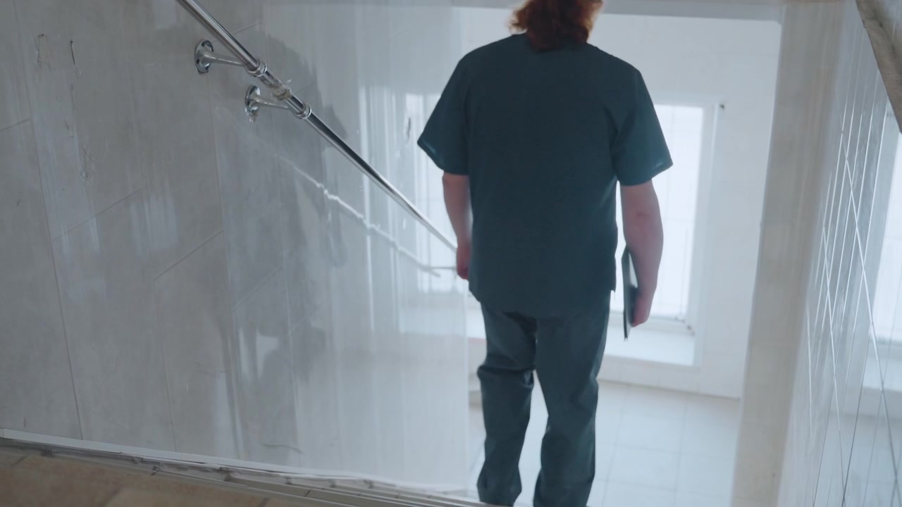 Paramedico che scende una scala piastrellata poco illuminata, vista posteriore di un'infermiera in divisa che porta una cartella clinica, lenta discesa verso il pianerottolo di un edificio residenziale, umore tranquillo e postura riflessiva dopo la visita a domicilio.
