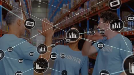 Animation-of-network-of-icons-over-three-diverse-volunteers-high-fiving-each-other-at-warehouse