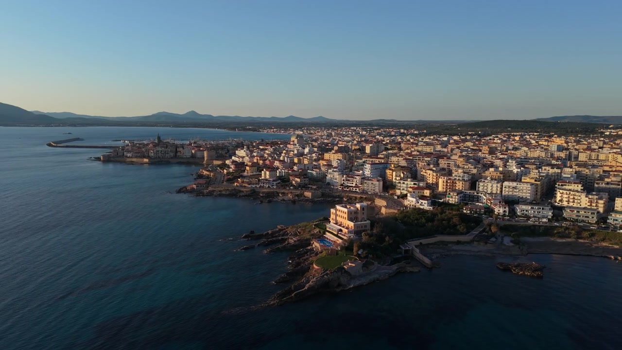 Isolati mediterranei costeggiano il lungomare roccioso mentre la telecamera scorre lungo la costa di Alghero nella calda luce della sera.