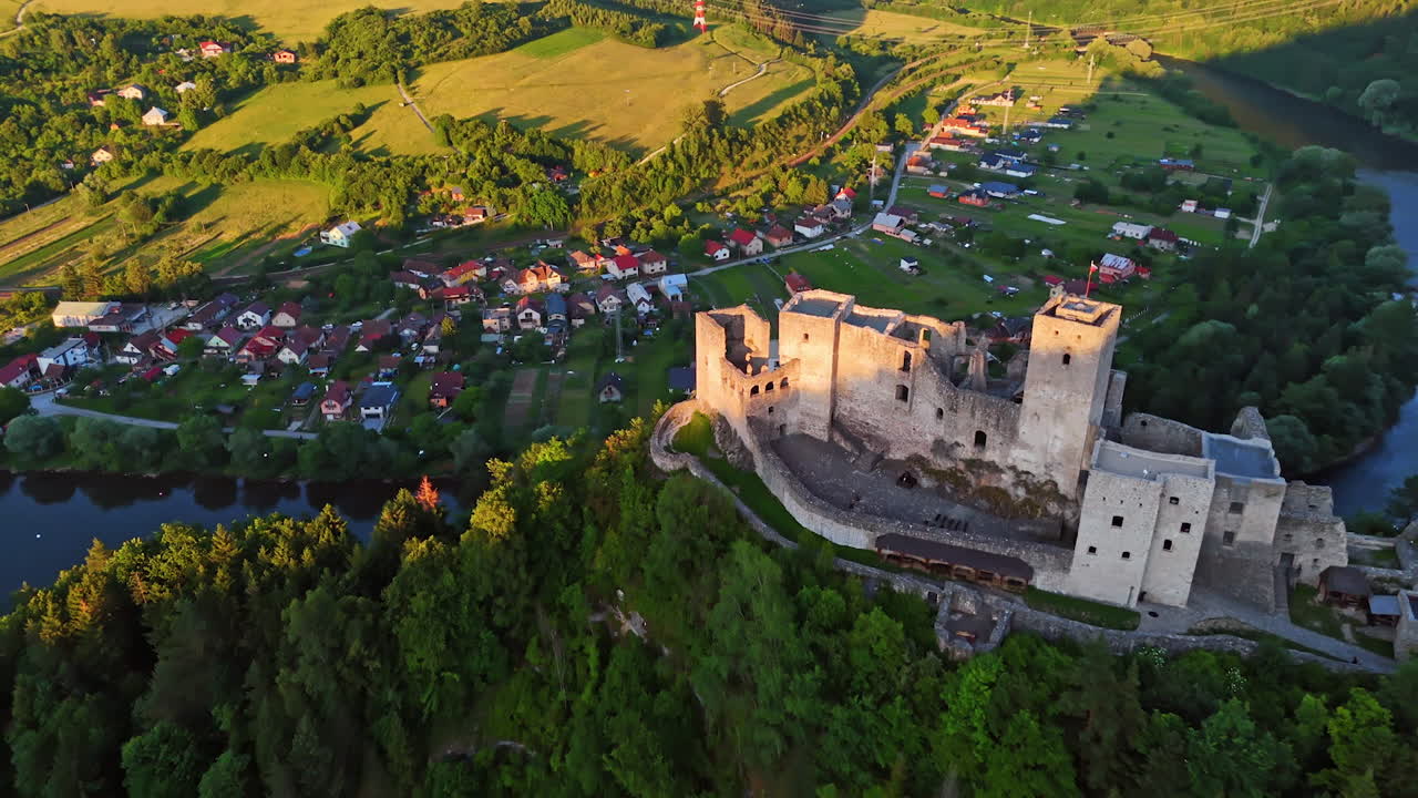 Ruins of a historic castle by the river. Aerial view of ancient castle ruins overlooking a peaceful river and a nearby village during a bright sunny day