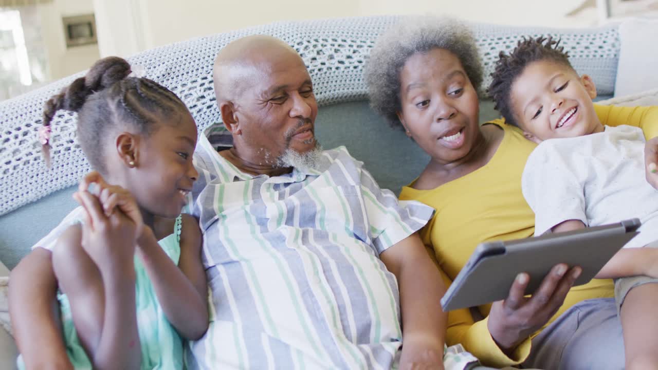 Happy african american grandparents, grandson and granddaughter on couch using tablet, slow motion