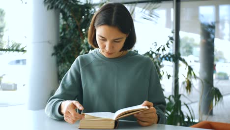 young attractive female student reads a book and ponders what she has read while sitting at a table in a public library. close-up portrait of a young cheerful female student, she is looking for the necessary information in the book, getting ready for the