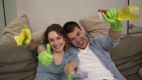 Portrait-of-a-young,-cheerful-couple-in-green-rubber-gloves.-The-family-finishing-the-house-cleaning-together.-Happy-tired-team,-sitting-on-a-couch,-looking-to-the-camera,-smiling,-thumbs-up