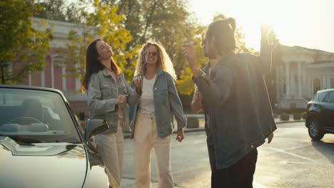 un grupo de jóvenes elegantes de pie cerca del coche, divertirse y bailar en el fondo de un día soleado