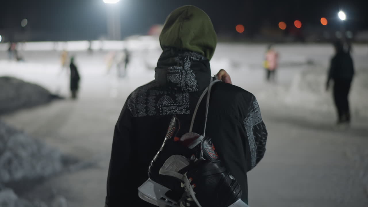 back view of person walking on snow covered rink at night wearing hooded jacket with skate boot slung over shoulder as distant figures and bright lights glow softly across snowy background