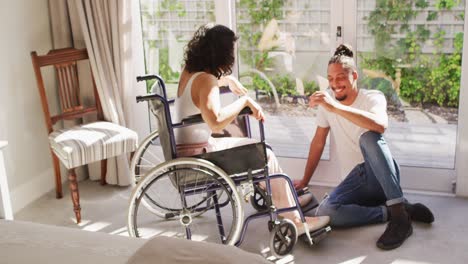 mujer biracial feliz en silla de ruedas y compañero masculino sonriente hablando en la sala de estar soleada