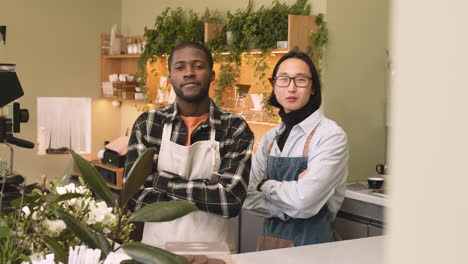 two multiethnic waiters with crossed arms looking at camera while standing behind counter in a coffee shop 1