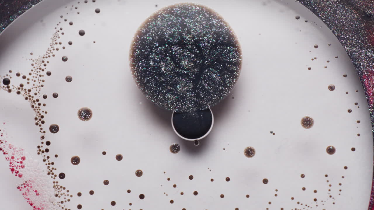Macro view of vibrating black glitter ink in a white liquid, surrounded by red and white ink droplets. The ink pulses and swirls intensely