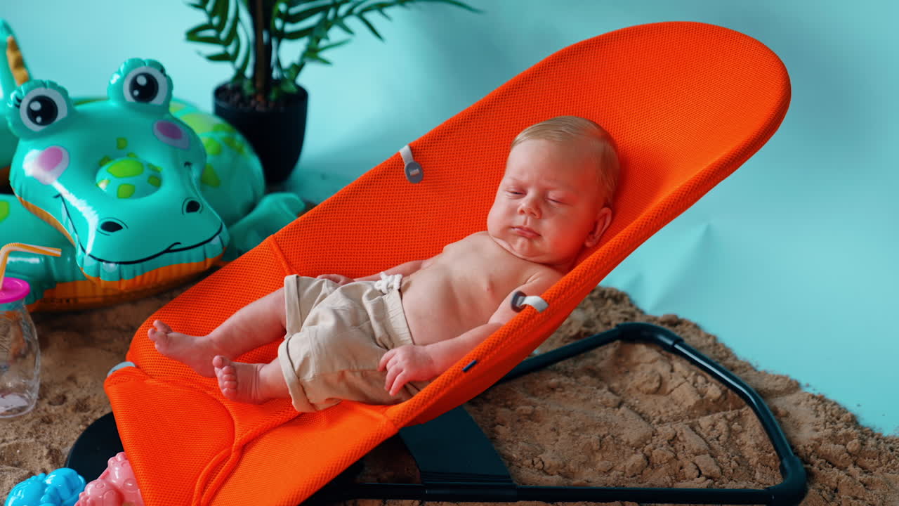 Blond tiny baby boy lying in the orange chair. Cute child resting at the improvised beach.