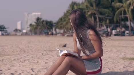 young woman with dark hair sits on sandy beach and reads