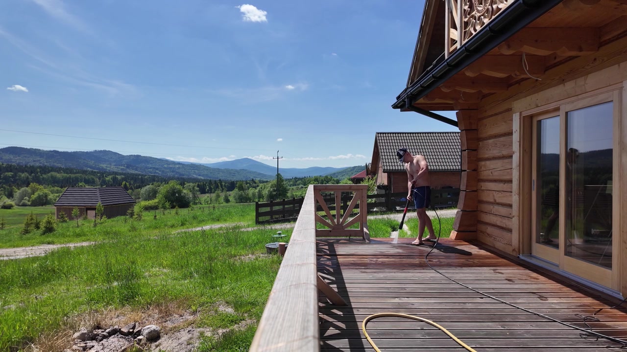 Uomo a torso nudo che indossa pantaloncini blu e un berretto lava una terrazza in legno con un'idropulitrice presso una baita sotto un cielo estivo di un blu brillante.