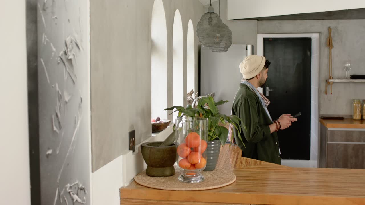 Man leaning on countertop in kitchen is scrolling and tapping phone under windows in daylight