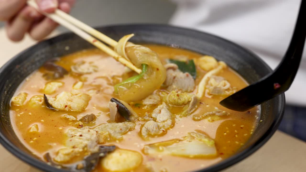 Hand uses chopsticks and spoon to lift vegetables from steaming spicy hotpot soup, bright lighting
