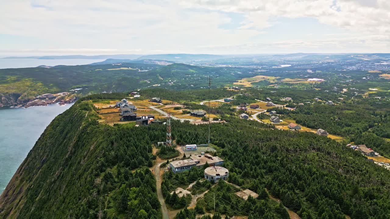 A drone soars above Red Cliff's inland plateau, showing a tall communications tower rising over rolling fields, open meadows and forested hills under a bright Newfoundland sky
