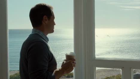 man looking out a window at the ocean, holding a coffee cup