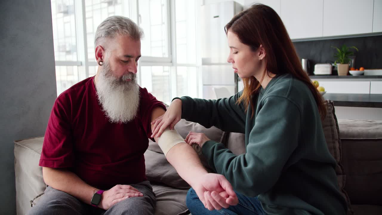 una chica morena en un suéter verde venda su mano con un vendaje a su padre anciano con cabello gris y una barba exuberante en una camiseta roja mientras está sentada en un sofá marrón en un apartamento moderno