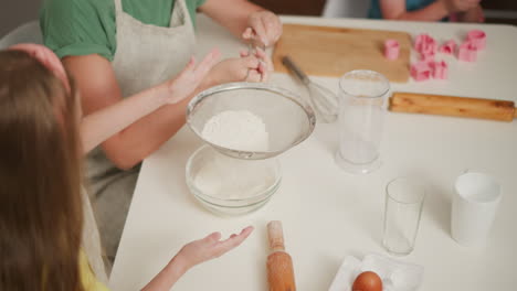 girl child taps sieve with left hand while instructor holds handle to sift flour into bowl, partial view of another child playing in background with baking tools on table in cozy indoor kitchen