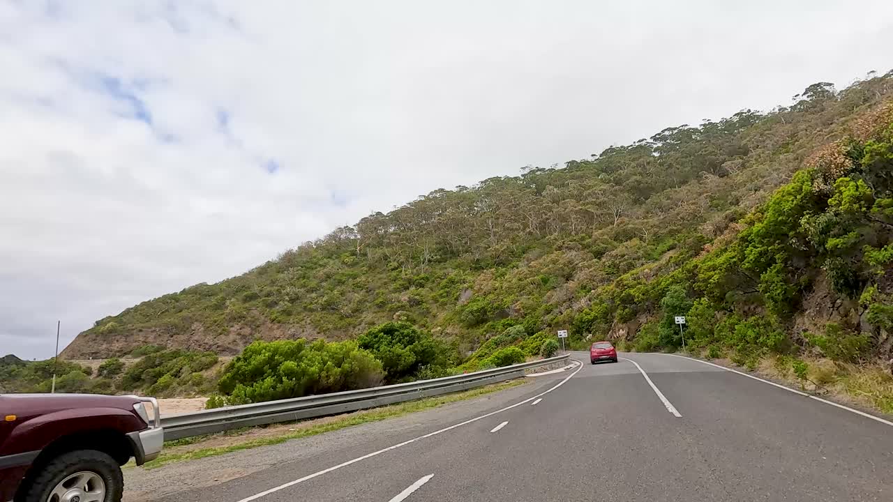 A car travels along the winding Great Ocean Road, surrounded by lush greenery and coastal views under a cloudy sky