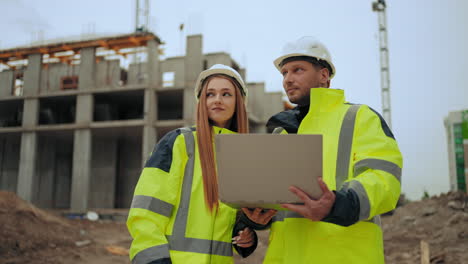 Dos-Ingenieros-Civiles-Están-Discutiendo-El-Proyecto-Y-El-Progreso-De-La-Construcción-De-Un-Edificio-Moderno-Al-Aire-Libre,-Hombre-Y-Mujer