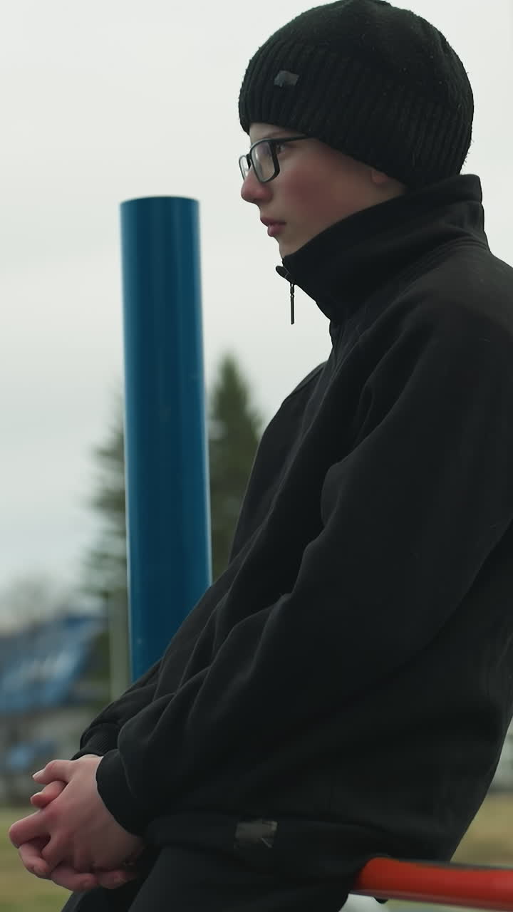 Side view of a young boy dressed in a black outfit and glasses, sitting thoughtfully on a red iron bar, with a blur view of people in the in the field