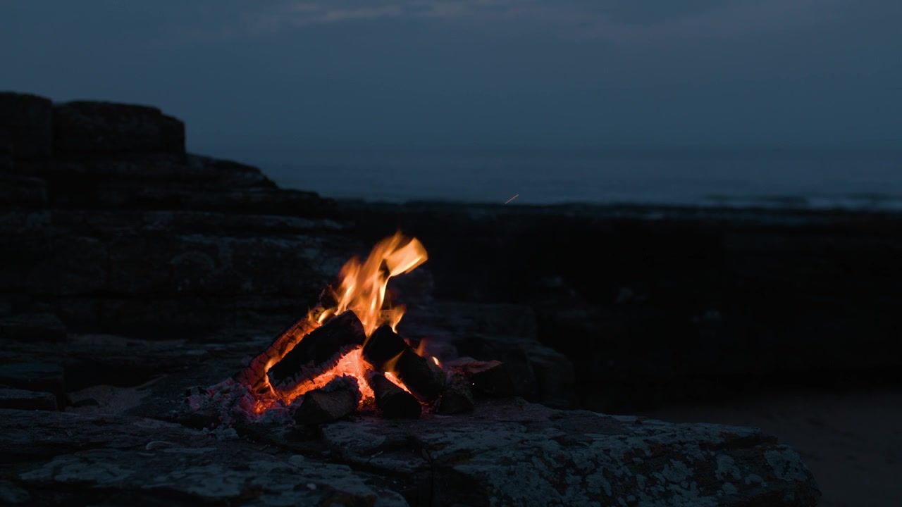 Fiamme calde e braci ardenti illuminano le rocce mentre la notte cala su una costa tranquilla, creando una scena marittima pacifica e suggestiva.