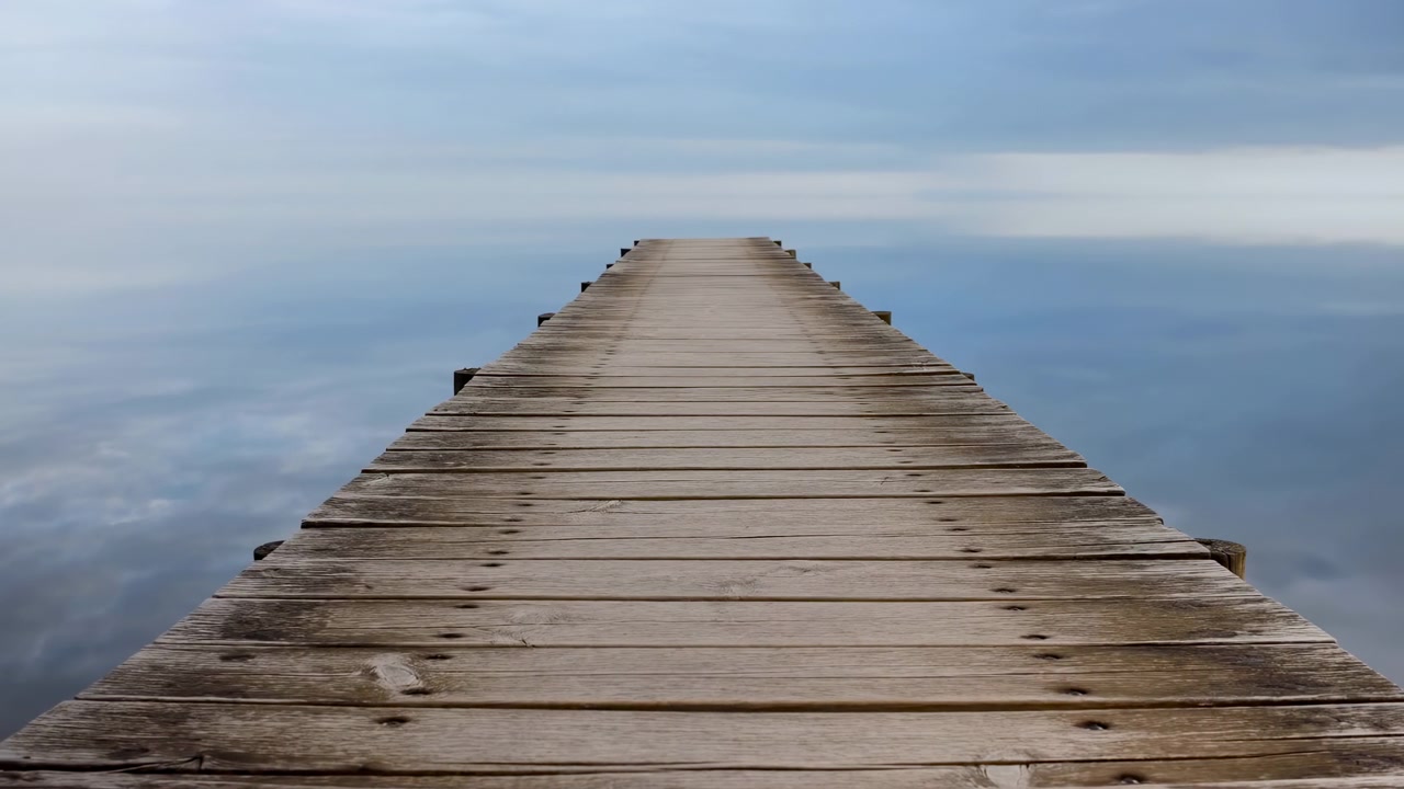 Pontile di legno che si estende su un lago calmo all'alba, assi consumate che conducono verso un orizzonte nebbioso, cielo dai colori pastello tenui.