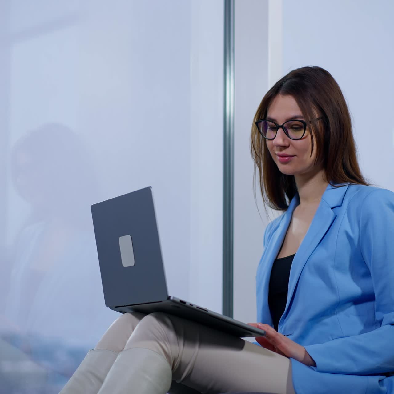 Relaxed positive brunette lady in blue jacket works on computer. Woman is typing on laptop sitting on a soft chair. Low angle view
